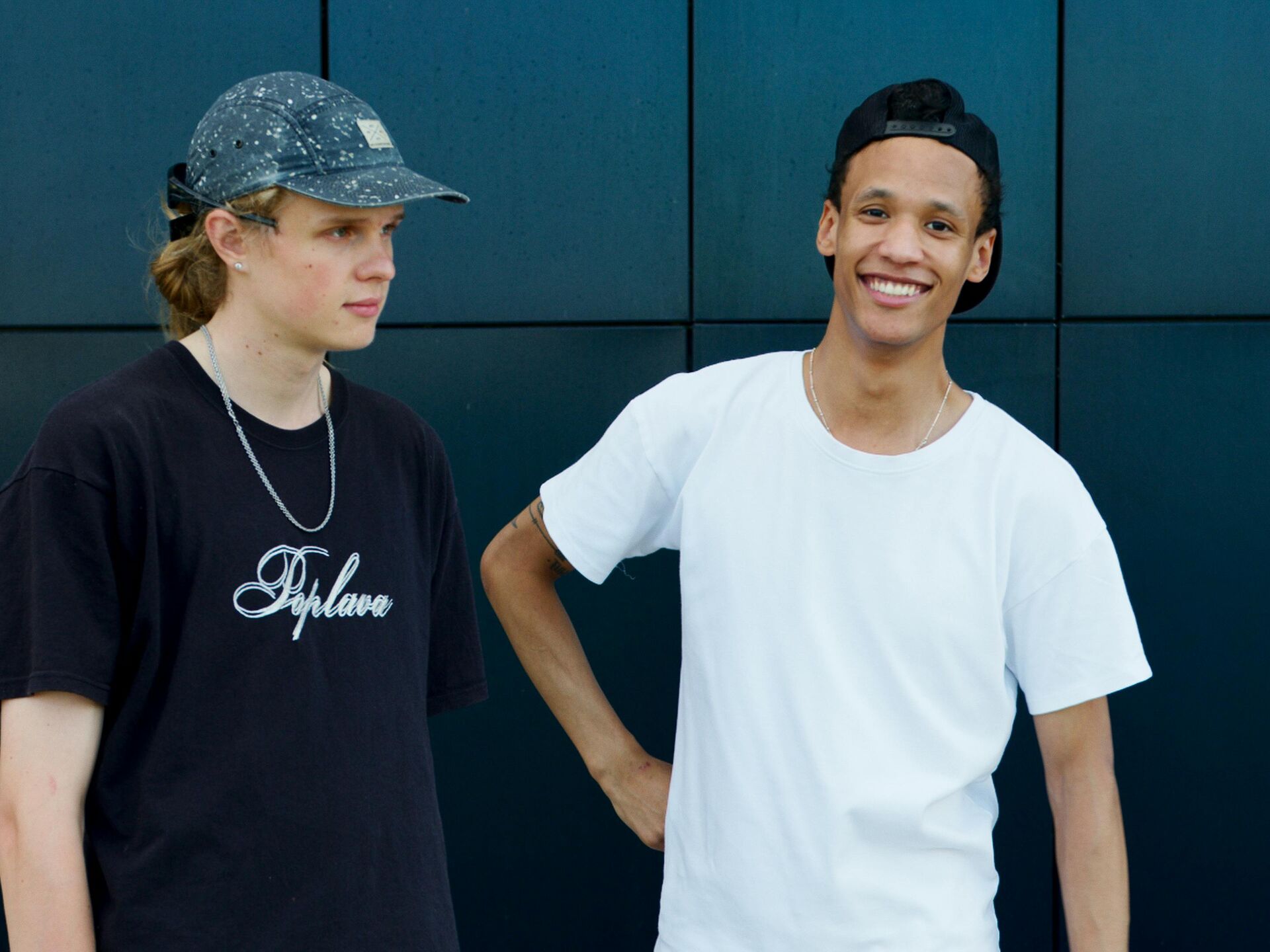Zwei junge Männer stehen vor einer dunklen Wand, einer blickt zur Seite, der andere lächelt in die Kamera.