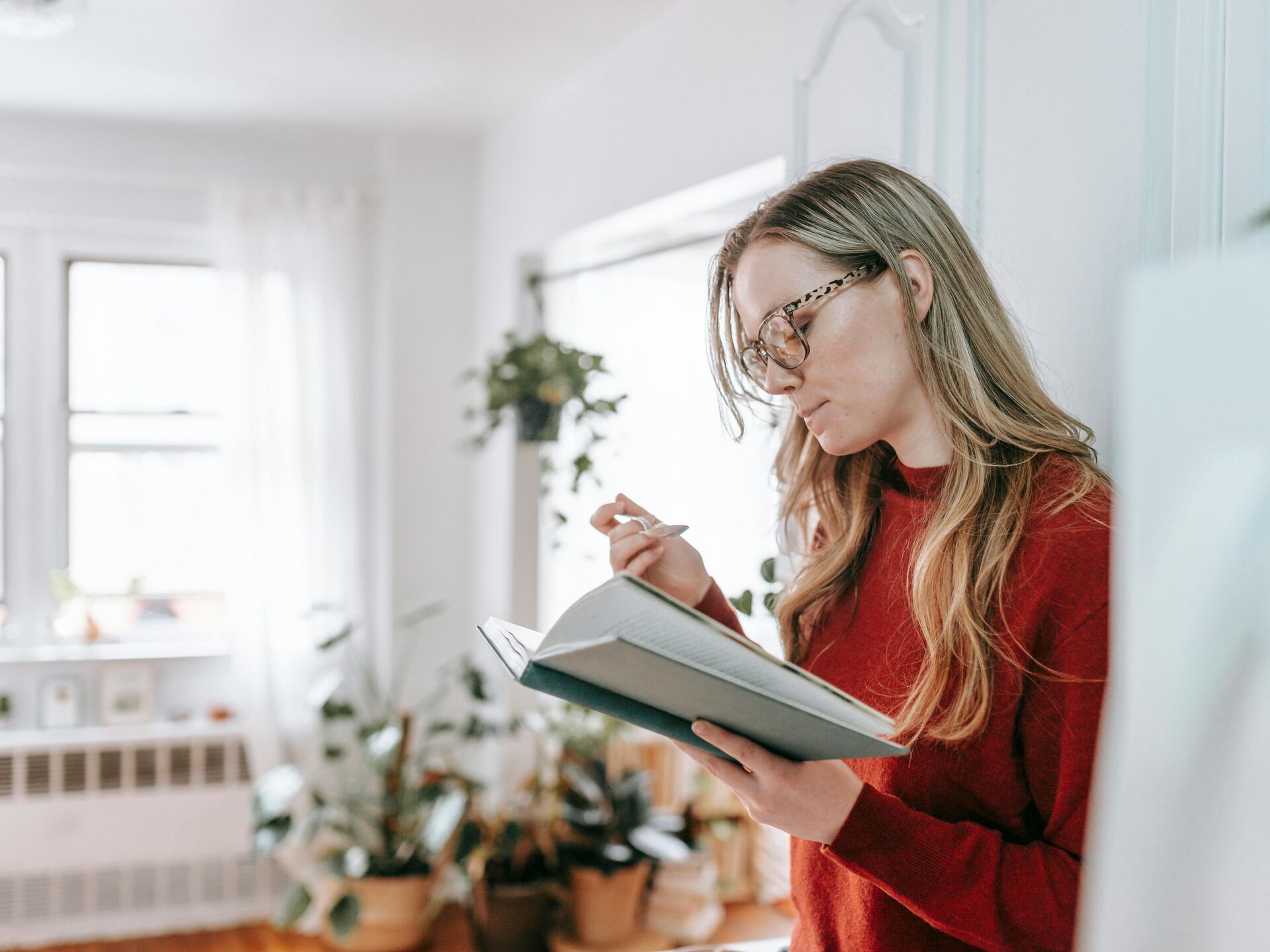Frau steht in einem hellen Raum mit vielen Pflanzen und liest konzentriert in einem geöffneten Notizbuch.