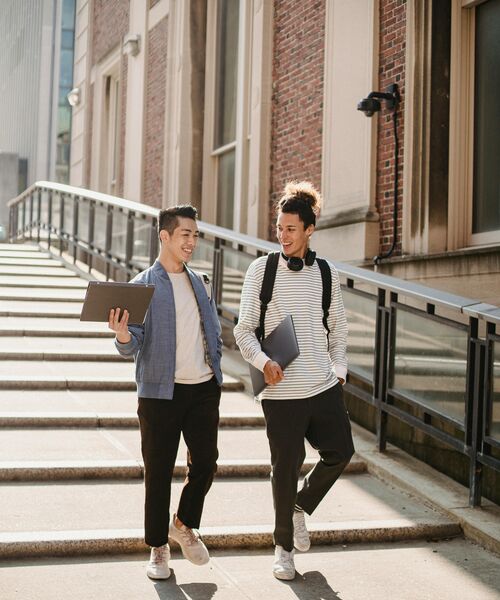 Zwei Studierende gehen lachend eine Treppe hinunter und unterhalten sich, jeweils mit Laptop in der Hand.