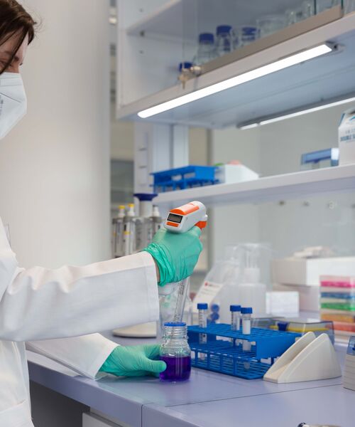 Laborperson mit Schutzmaske und Handschuhen arbeitet an einer Pipette und füllt eine violette Flüssigkeit in ein Gefäß.
