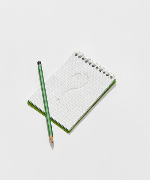 Notizblock mit karierten Seiten und gezeichnetem Fragezeichen, daneben ein grüner Bleistift.