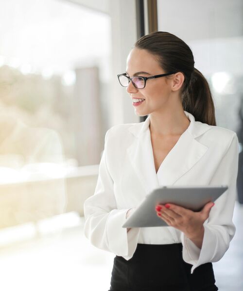 Eine lächelnde Frau in weißem Blazer hält ein Tablet und blickt zur Seite in einen hell erleuchteten Raum.