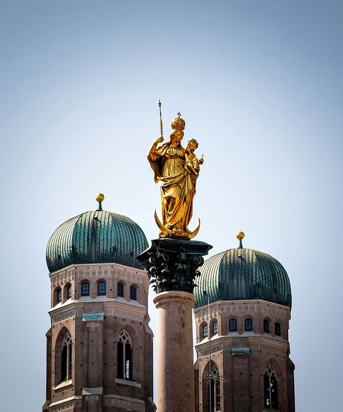 Goldene Marienstatue auf der Mariensäule vor den markanten Zwiebeltürmen der Münchner Frauenkirche.