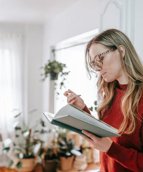 Frau steht in einem hellen Raum mit vielen Pflanzen und liest konzentriert in einem geöffneten Notizbuch.