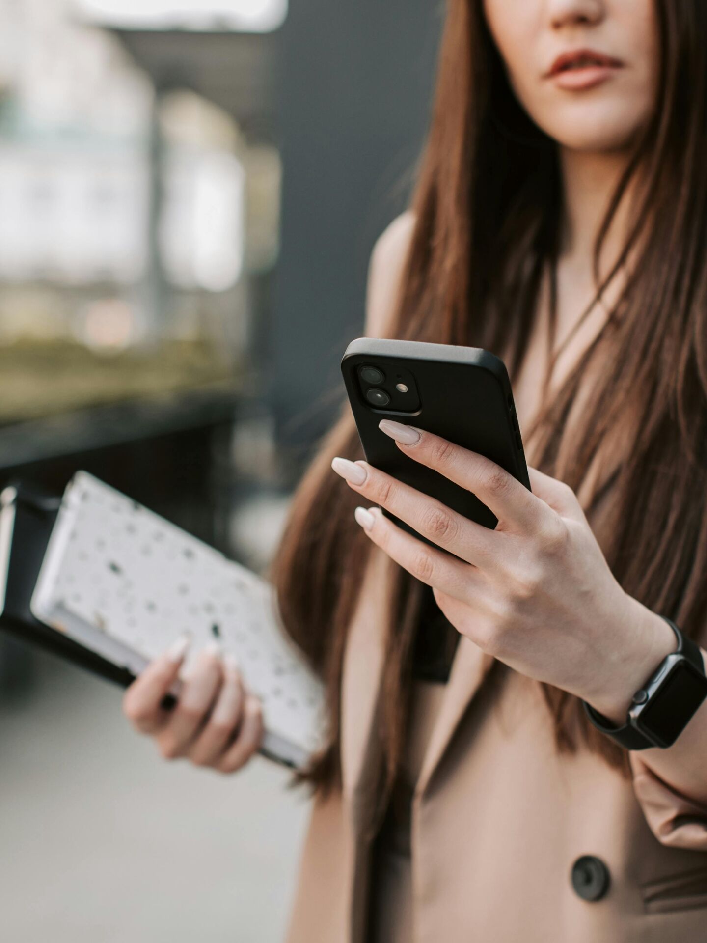 Frau hält ein Smartphone in der Hand und trägt Unterlagen sowie eine Smartwatch, Fokus auf Hände und Gerät.