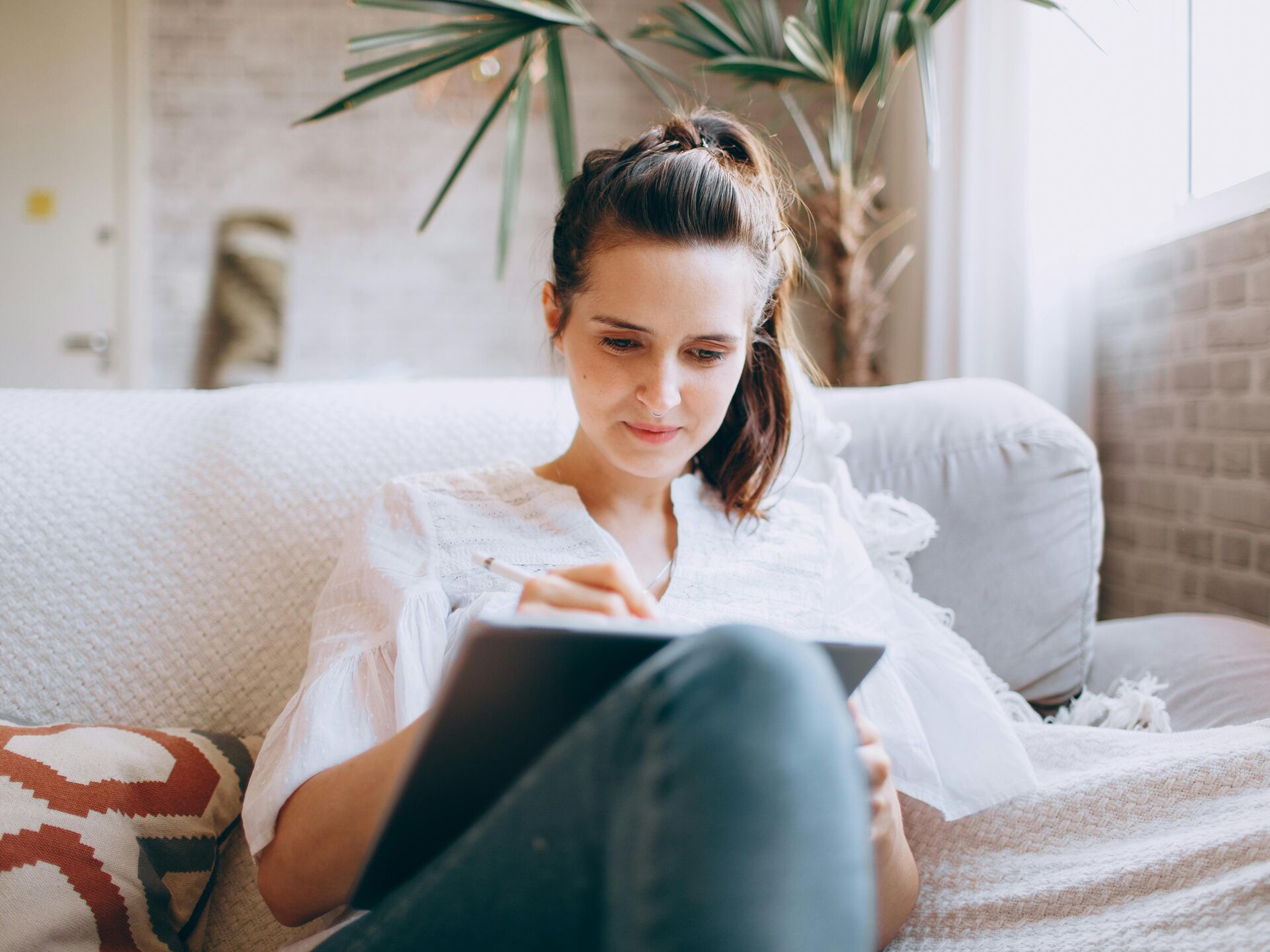 Frau sitzt entspannt auf einem Sofa und arbeitet konzentriert mit einem Tablet und Stylus.