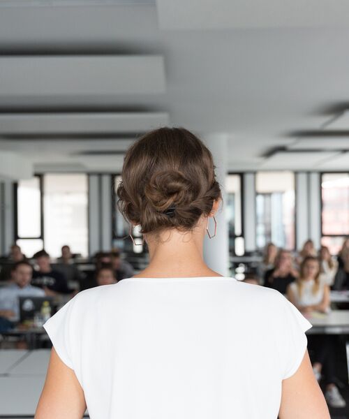Frau steht mit dem Rücken zur Kamera vor einer Gruppe von Menschen in einem Seminar- oder Vortragsraum.