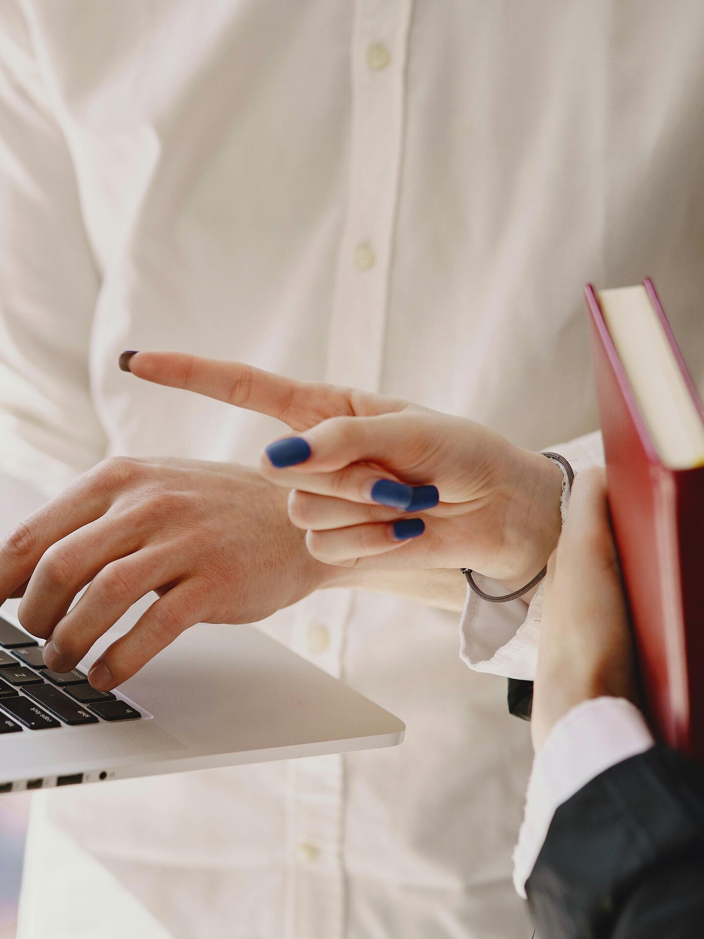 Person zeigt mit der Hand auf den Bildschirm eines Laptops, während eine andere Person daneben ein Buch hält.