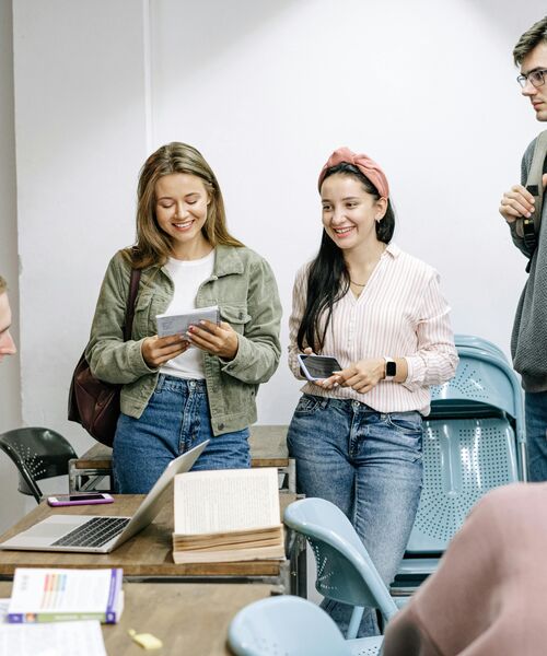 Gruppe junger Menschen steht lächelnd in einem Raum zusammen und betrachtet Unterlagen; im Vordergrund liegt ein Laptop auf einem Tisch.