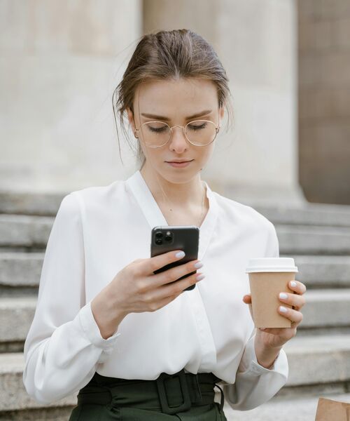 Eine junge Frau steht draußen auf Treppenstufen, hält einen Kaffeebecher und schaut konzentriert auf ihr Smartphone.