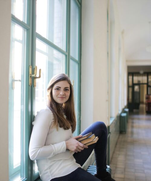 Eine junge Frau sitzt entspannt auf einer Fensterbank in einem hellen Flur und hält mehrere Bücher in den Händen.