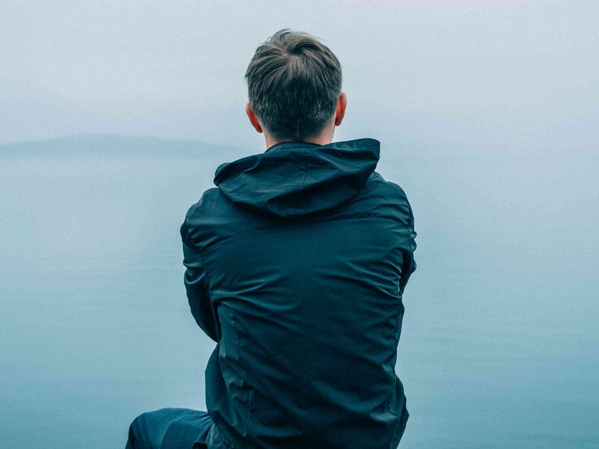 Person sitzt mit dem Rücken zur Kamera und blickt über ein ruhiges Gewässer.