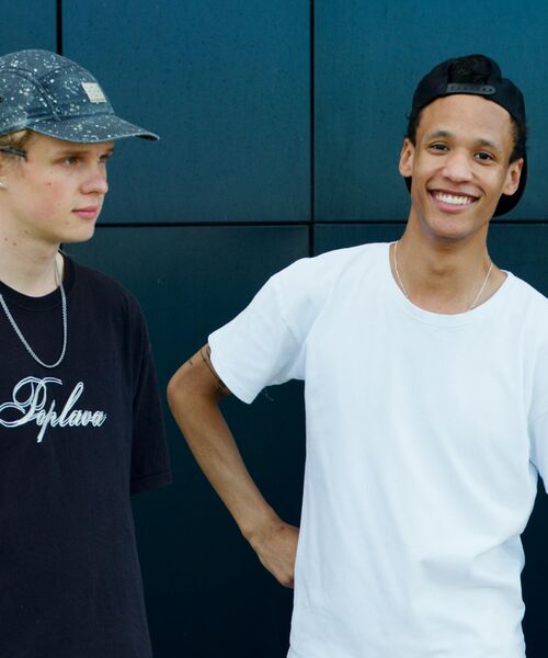 Zwei junge Männer stehen vor einer dunklen Wand, einer blickt zur Seite, der andere lächelt in die Kamera.