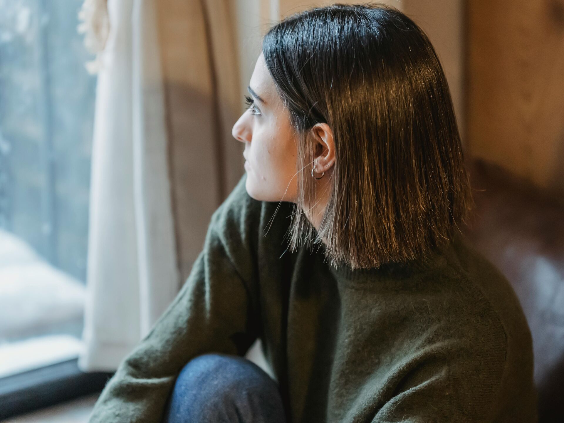 Person blickt nachdenklich aus dem Fenster, in gemütlicher Umgebung sitzend.