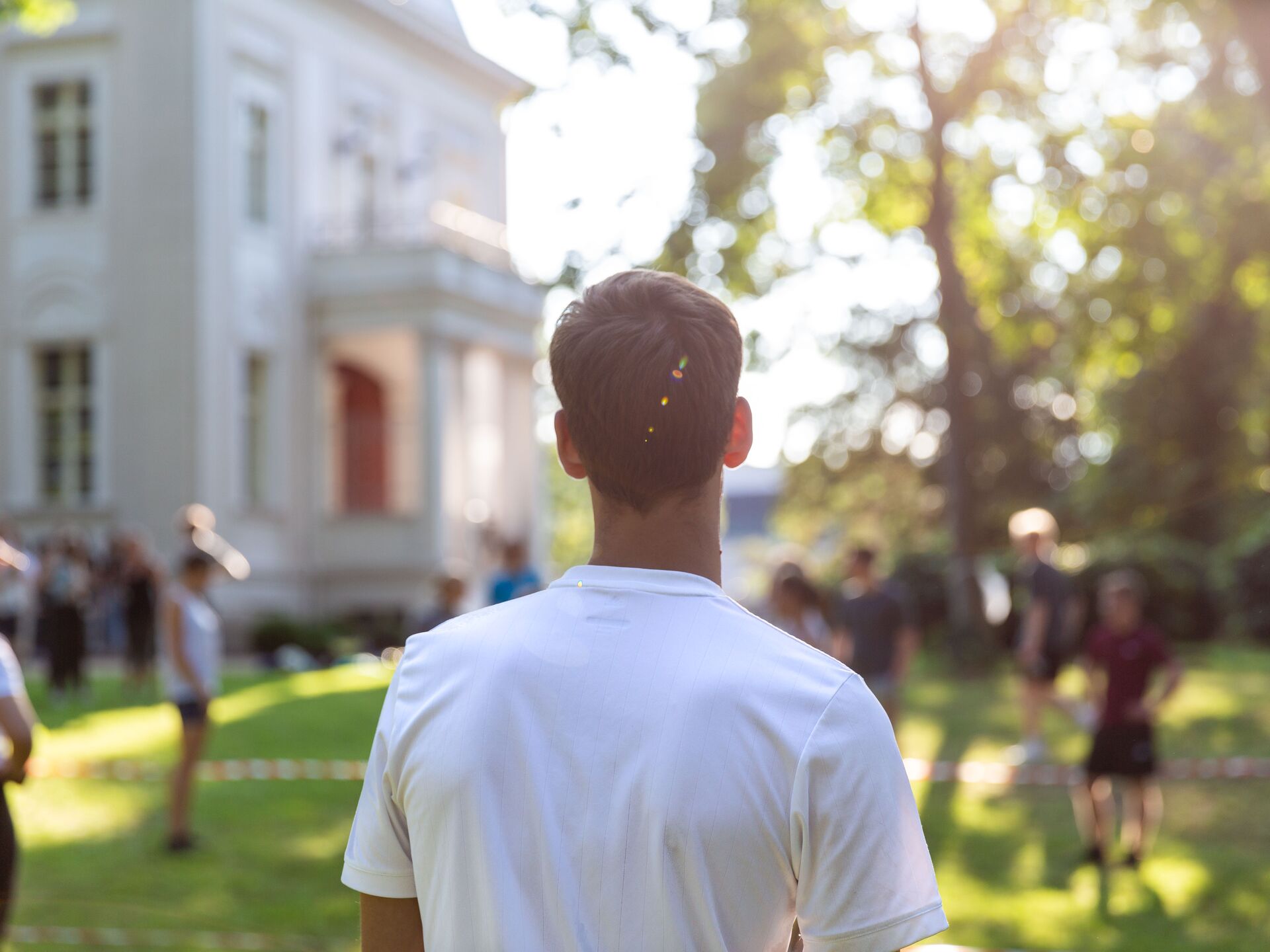 Person steht mit dem Rücken zur Kamera auf einer sonnigen Grünfläche vor einem hellen Gebäude.