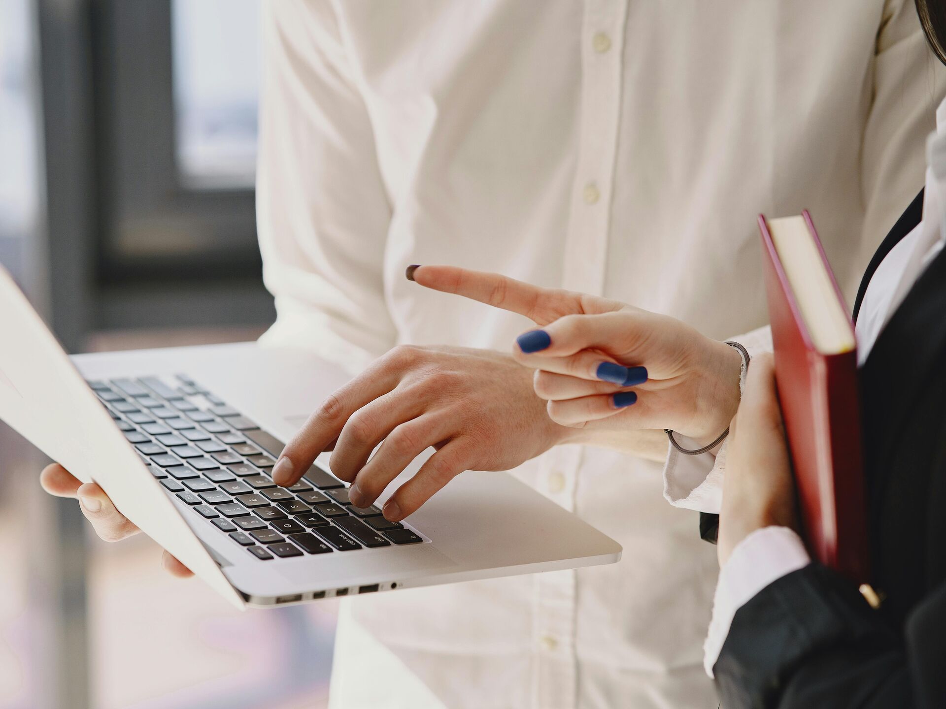 Person zeigt mit der Hand auf den Bildschirm eines Laptops, während eine andere Person daneben ein Buch hält.