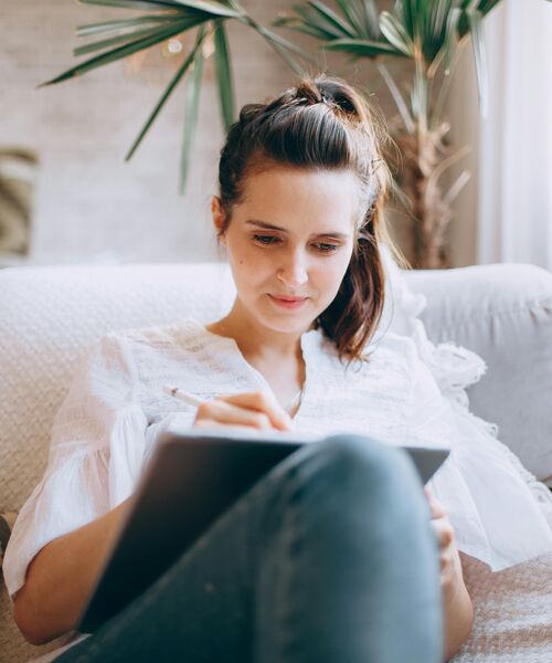Frau sitzt entspannt auf einem Sofa und arbeitet konzentriert mit einem Tablet und Stylus.