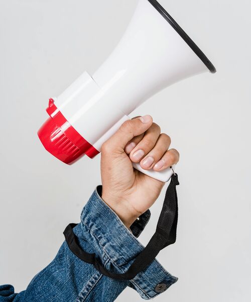 Hand in Jeansjacke hält ein weiß-rotes Megafon vor hellem Hintergrund.