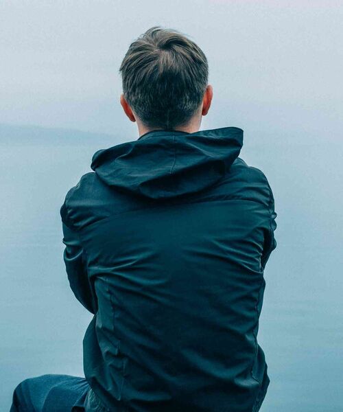 Person sitzt mit dem Rücken zur Kamera und blickt über ein ruhiges Gewässer.