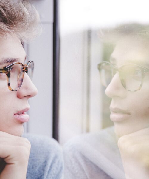 Person mit Brille blickt nachdenklich aus dem Fenster und sieht ihr Spiegelbild im Glas.