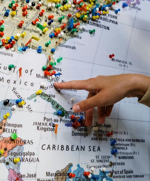 Hand zeigt auf eine Weltkarte mit vielen bunten Stecknadeln, die verschiedene Reiseziele markieren.