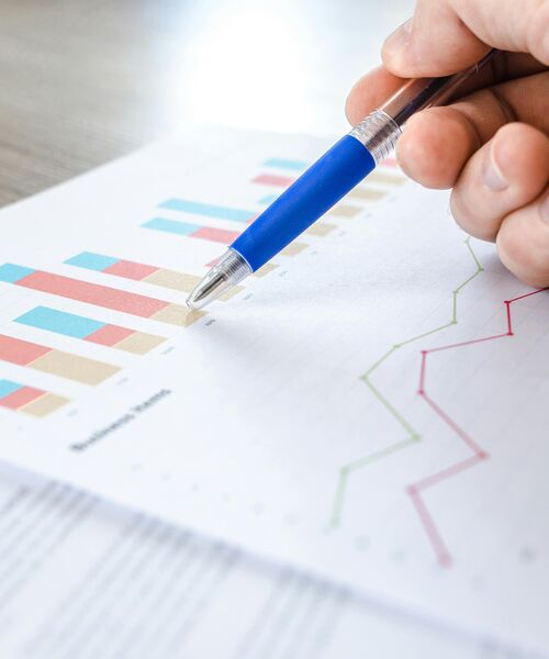 Nahaufnahme einer Hand mit Stift, die auf ein Blatt mit farbigen Diagrammen und Grafiken zeigt.