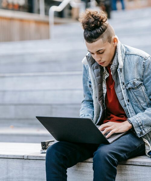 Junger Mann sitzt draußen auf Treppenstufen und arbeitet konzentriert an einem Laptop.