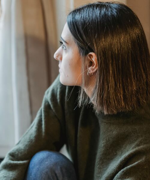 Person blickt nachdenklich aus dem Fenster, in gemütlicher Umgebung sitzend.