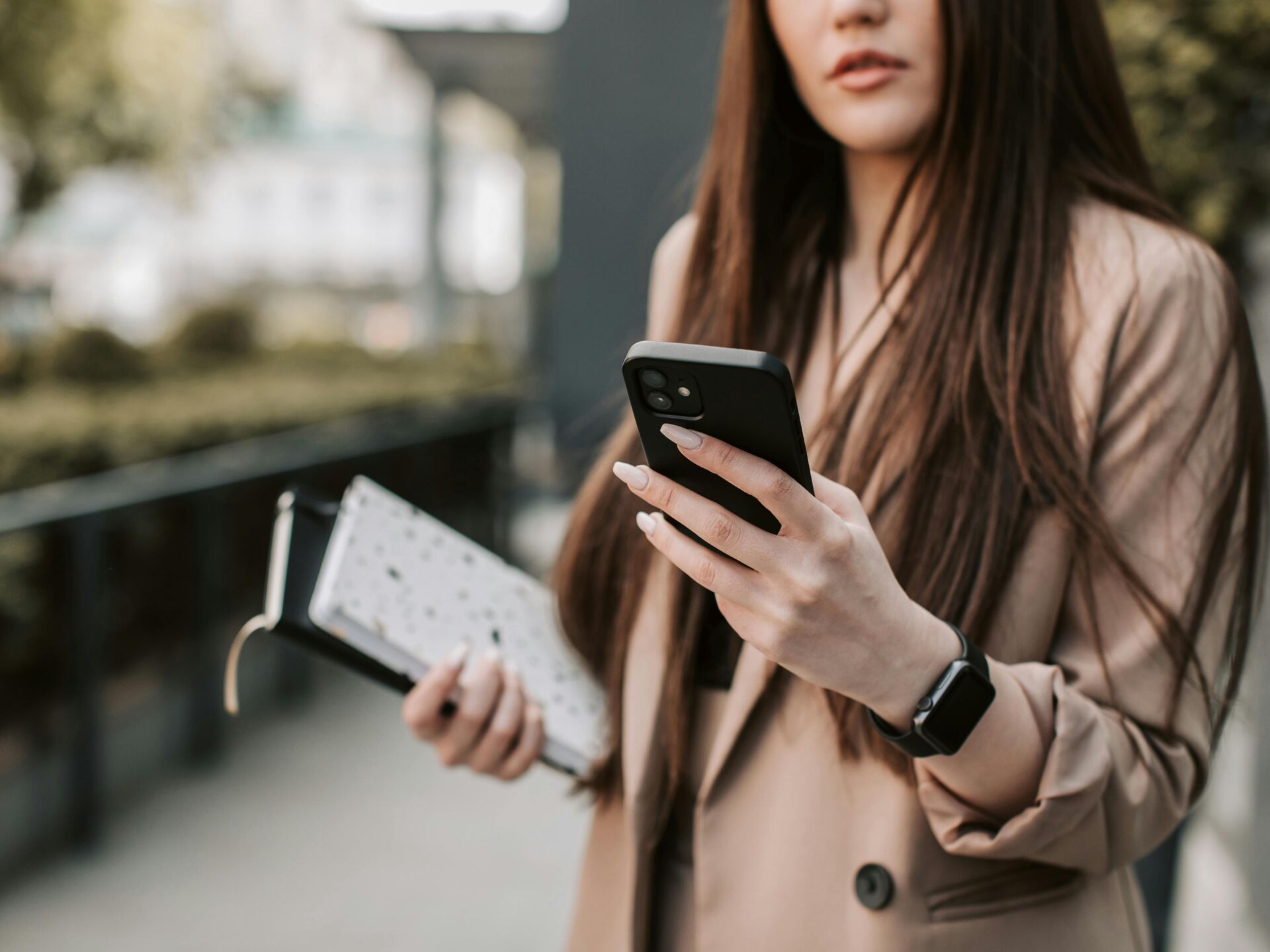 Frau hält ein Smartphone in der Hand und trägt Unterlagen sowie eine Smartwatch, Fokus auf Hände und Gerät.