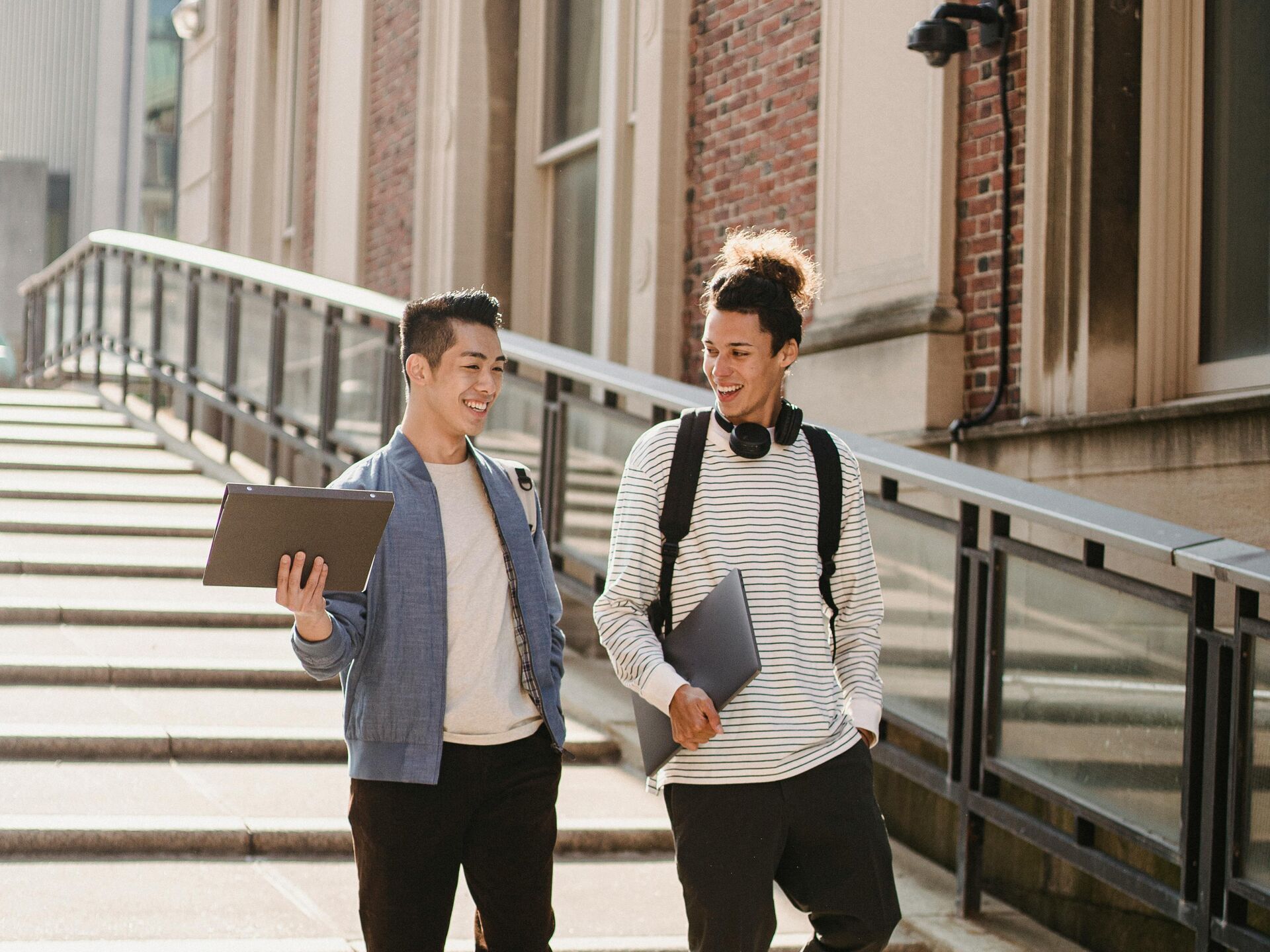 Zwei Studierende gehen lachend eine Treppe hinunter und unterhalten sich, jeweils mit Laptop in der Hand.