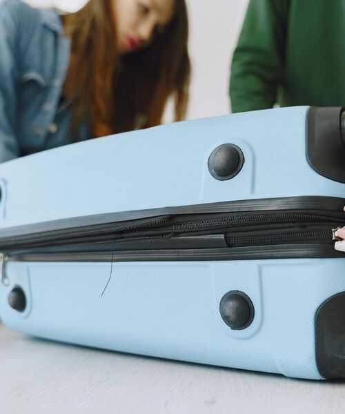 Hellblauer Koffer in Nahaufnahme, im Hintergrund zwei unscharfe Personen – Symbol für Reisen oder Aufbruch.