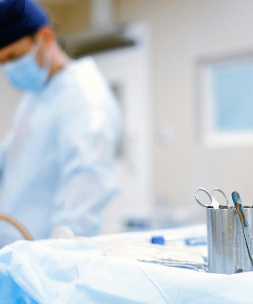Sterile Operationsumgebung mit chirurgischen Instrumenten im Vordergrund und einer Person im OP-Kittel und Mundschutz im Hintergrund.