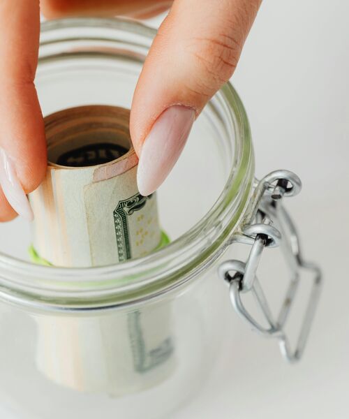 Hand legt eine zusammengerollte Geldscheinnote in ein Glasgefäß.