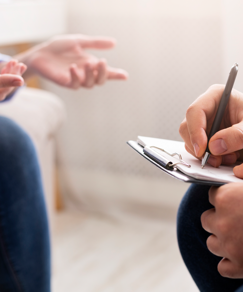 Gesprächsszene zwischen zwei Personen, eine Person macht Notizen auf einem Klemmbrett.