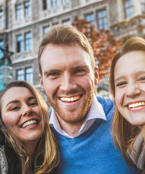 Drei junge Erwachsene stehen dicht zusammen draußen und lächeln fröhlich in die Kamera vor einem historischen Gebäude.