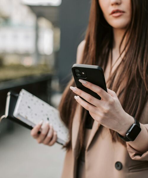 Frau hält ein Smartphone in der Hand und trägt Unterlagen sowie eine Smartwatch, Fokus auf Hände und Gerät.