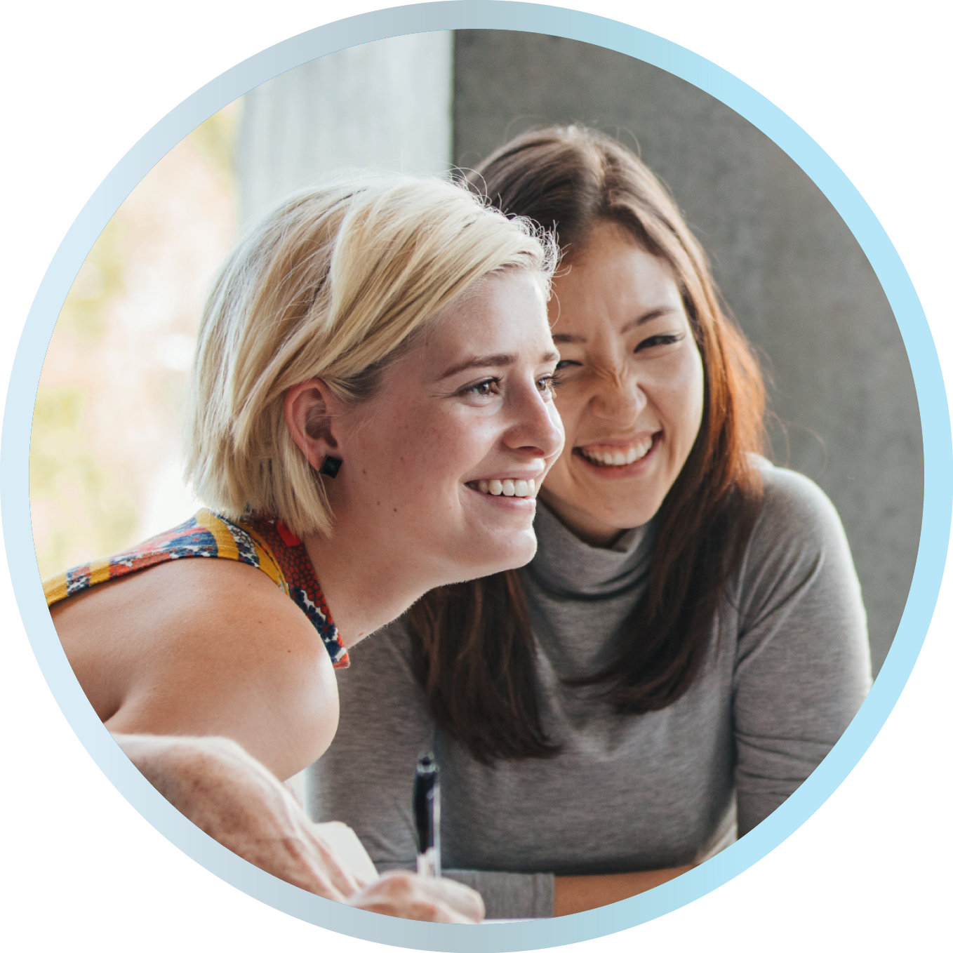 Zwei lächelnde Frauen sitzen nebeneinander und arbeiten gemeinsam an einem Tisch.